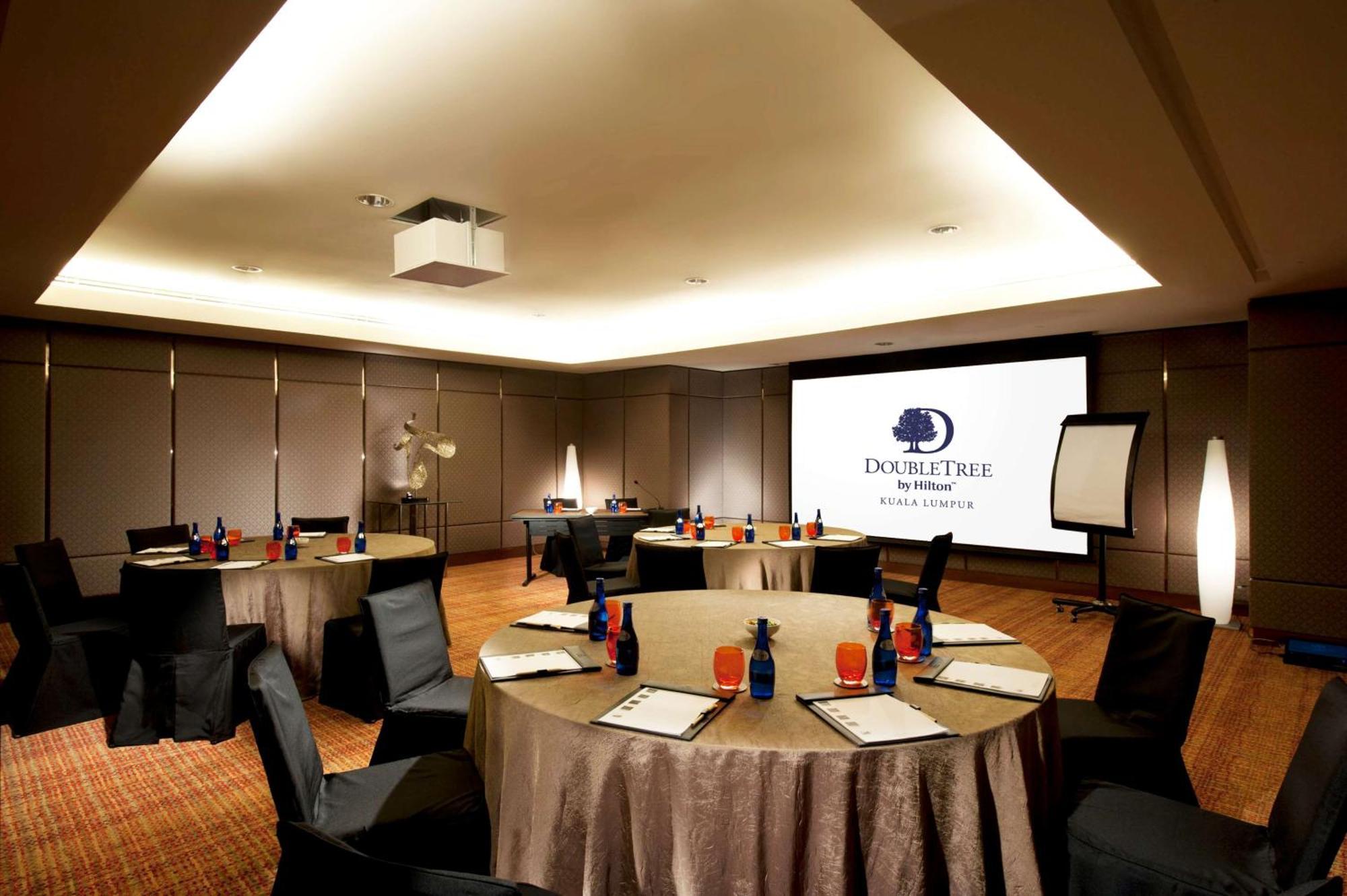 Готель Doubletree By Hilton Куала-Лумпур Екстер'єр фото The photo shows a conference room set up for a meeting or event. There are several round tables with beige tablecloths, each adorned with blue and red decorations, likely drinks and supplies. Black chairs are placed around the tables. On one wall, th
